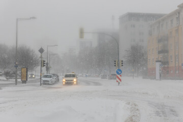 Severe snow storm hits Magdeburg city intersection cars taxi moving slowly whiteout conditions low visibility. Snow covered road traffic lights urban buildings weather disruption danger winter