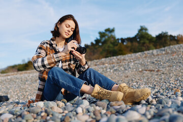 Woman sitting on a pebble beach, outdoors, holding a camera for photography, wearing a plaid shirt...