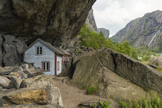 View of a quaint, weathered house nestled precariously beneath a colossal rock overhang, juxtaposed against the rugged, mountainous terrain, Helleren, Jossingfjord, Norway.