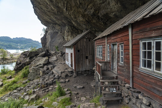 View of weathered wooden houses nestled beneath a colossal rocky overhang, where rugged terrain meets serene waters, creating a timeless scene, Helleren, Jossingfjord, Norway.