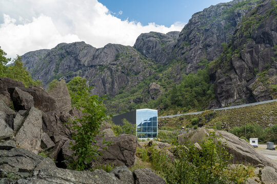 View of a modern glass structure stands in stark contrast to the rugged, towering mountains under a partly cloudy sky, Helleren, Jossingfjord, Norway.