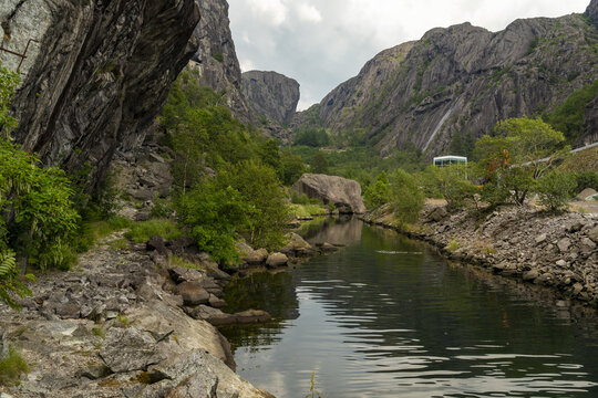 View of a serene canal reflecting the rugged cliffs and lush greenery under an overcast sky, creating a dramatic contrast of light and shadow, Helleren, Jossingfjord, Norway.