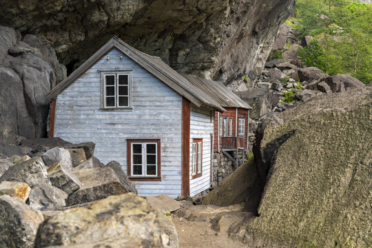 View of a weathered, light-blue wooden house nestled tightly beneath a massive, rocky overhang, a testament to nature's embrace, Helleren, Jossingfjord, Norway.