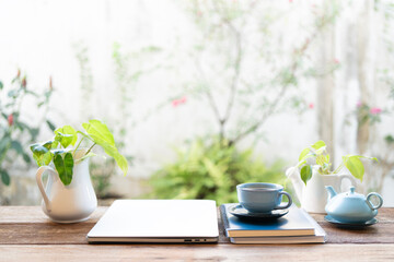 Folded laptop and blue tea set and plant pot on wooden table with backyard view