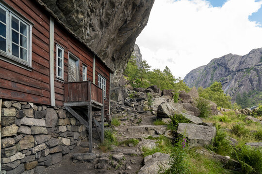 View of a red wooden house nestled under a massive overhanging rock formation, standing in stark contrast to the rugged stone steps, Helleren, Jossingfjord, Norway.