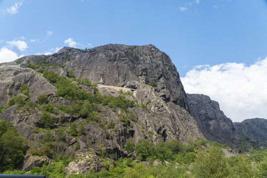 View of a towering, dark rocky mountain face with sparse green vegetation under a bright blue sky, a natural fortress of stone, Helleren, Jossingfjord, Norway.