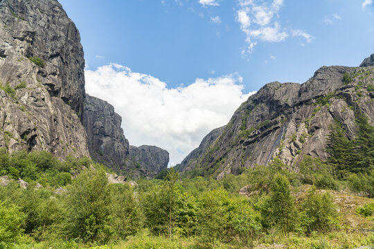 View of imposing granite cliffs embrace a verdant valley under a summer sky dotted with fluffy clouds, a serene tableau of nature's grandeur, Helleren, Jossingfjord, Norway.