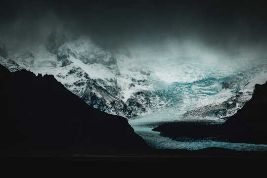 View of dark, brooding mountains frame a stunning glacial river winding its way through a stark, snow-dusted landscape under a cloudy sky, Vik, South Island, Iceland.