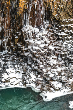 View of icy basalt columns cascading into the turquoise river below, a surreal landscape where winter's grip meets the vibrant heart of nature, East Fjords, Iceland.
