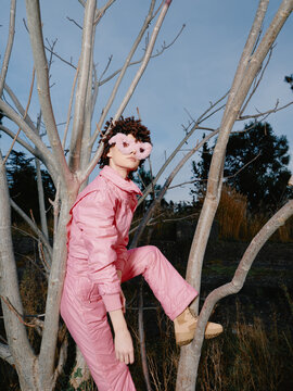 Pink fashion portrait among leafless branches, creative silhouette and whimsical vibe, a model in soft pink suit climbing a tree with floral glasses under clear sky