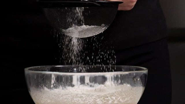 Baker measures and sifts flour into a large mixing bowl in a kitchen. Using a sifter to add flour into a large bowl for baking banana bread