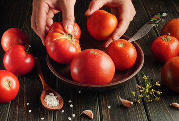 Hands are picking fresh tomatoes from a bowl placed on a dark wooden surface. Garlic cloves and salt sit nearby along with a knife, showing preparation for cooking
