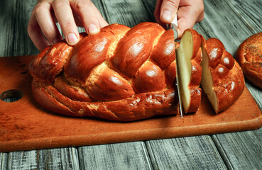 Hands are cutting a loaf of bread on a wooden board. The scene takes place in a kitchen where bread is being prepared for serving