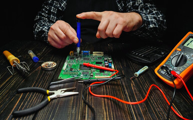 A person is focused on repairing a circuit board using various tools on a dark wooden table. Tools like pliers and a multimeter are nearby, showing an electronics repair scene