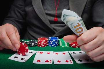 A man sits at a green felt-covered table covered with playing cards and stacks of poker chips. He holds a wad of cash. The scene takes place in a dimly lit casino