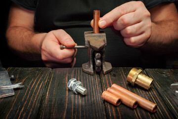 A person uses a tool to work on copper pipes and fittings on a wooden table in a workshop. Metal parts are spread out nearby as the work progresses in the evening light