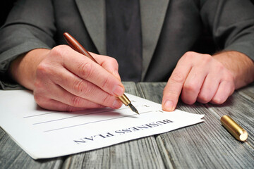A person is seated at a table, writing on a sheet of paper labeled business plan. The individual uses a fountain pen, focusing on the task with attention