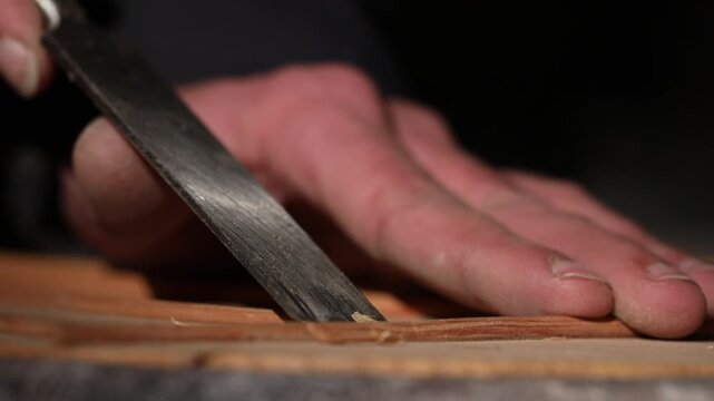 Close-up of a luthier's hands carefully chiseling wooden braces on a guitar soundboard. The scene highlights traditional craftsmanship and precision in a dimly lit instrument workshop.

