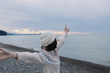 Woman on a pebbled beach with outstretched arms toward the sea at sunset, wearing a knit sweater and beanie, conveying freedom and calm.
