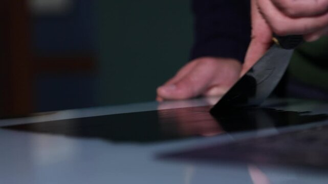 A close-up shot of an artist's hand using a palette knife to spread smooth black ink on a glass surface, preparing for a printmaking or linocut project.