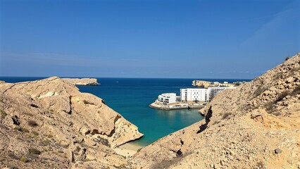 Quiet Bay Surrounded By Steep Rocky Cliffs, Sunlit Shore With Colorful Buildings And Tranquil Turquoise Waters, Calm Inlet Nestled Among Rugged Cliffs With Shimmering Sand And Peaceful Ambiance