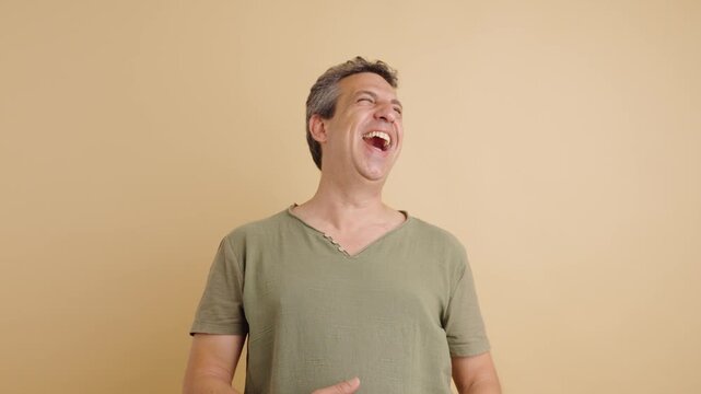 Happy middle-aged man pointing and laughing hysterically. Genuine amusement and positive emotions on display against a warm beige background