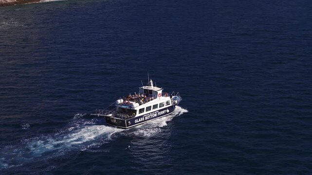 Aerial view shows GLASS BOTTOM FERRY with tourists off Gran Canaria, Atlantic waters, rocky coast, strong blues, high contrast, wake trails as the boat moves