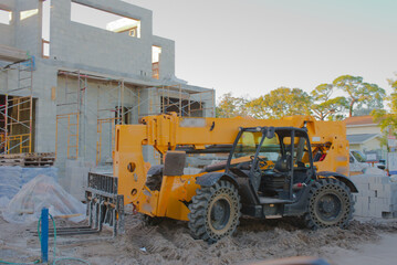 Obraz premium Construction Site With Modern Two-Story Block Building, Scaffolding, and Heavy Machinery At Work. Large yellow construction crane. Palms and cleared ground emphasize a developing site with active work