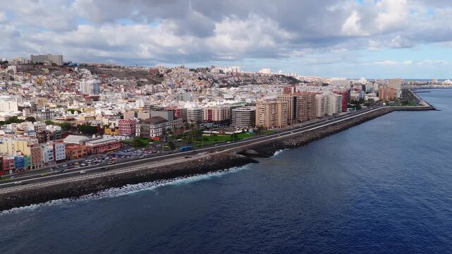 Aerial daylight view of Las Palmas de Gran Canaria coastline, seawall, Avenida Maritima, twin tower Catedral de Santa Ana, pastel facades, buses and cars, Atlantic waves