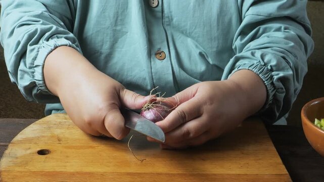 A woman's hands peel a red onion with a knife on a cutting board on the table and place it on a plate. Medium shot.