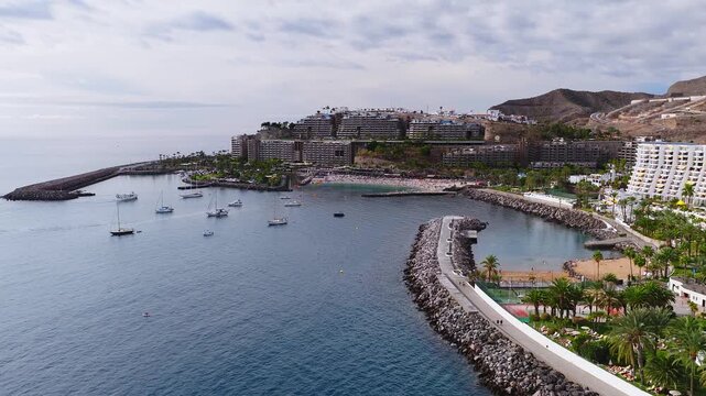 Aerial view shows curved breakwaters, anchored sailboats, sandy beach, terraced hillside hotels, kayaks, palm promenade, pools, courts, and arid volcanic hills.