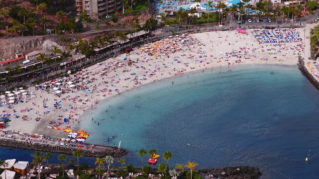 Aerial view of Playa de Amadores in Gran Canaria with palm lined crescent beach, rock jetties, stone promenade, hotels, pools, swimmers, umbrellas, and yachts.