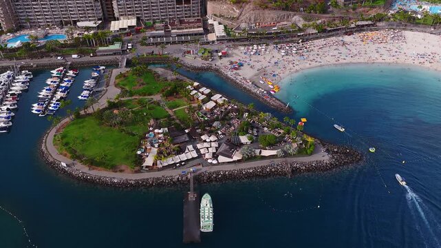 Aerial view of Playa de Amadores, Gran Canaria, shows hotels, marina, ferry pier, sunbeds, umbrellas, swimmers, small boats, breakwaters, and vivid blue bay in daytime