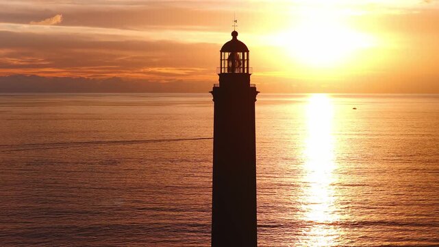 Aerial 4K shows Faro de Maspalomas at sunset in Gran Canaria. A boat leaves a thin wake as Teide silhouettes on the horizon and the sun reflects across the Atlantic.
