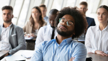 Diverse audience of professionals seated in a conference room, individual resting eyes appearing disengaged, business training or seminar atmosphere, group focus differing levels of attention