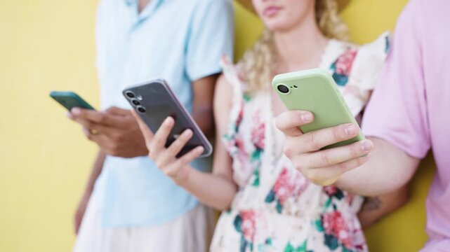 Young diverse group of people standing together against a yellow wall, each person focused on their own smartphone, scrolling and typing