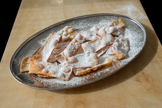 Tray of homemade chiacchiere with icing sugar leaning on a wooden board. Italian sweet dessert deep fried known also as sprelle, crostoli, angel wings, frappe, bugie or cenci. Carnival dessert