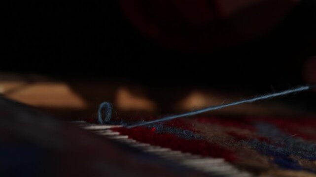 Close-up of elderly hands weaving a traditional rug on a loom. The artisan carefully threads red yarn through the warp strings in a dimly lit workshop setting.

