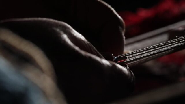 Close-up of elderly hands weaving a traditional rug on a loom. The artisan carefully threads red yarn through the warp strings in a dimly lit workshop setting.

