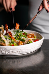 A person is about to eat a delicious Caesar salad with large grilled shrimp, green lettuce, parmesan cheese, and croutons in a white bowl.