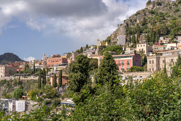 Obraz premium Picturesque hillside town in Sicily with colorful historic buildings cascading along steep slopes, surrounded by lush Mediterranean vegetation. The architecture blends harmoniously with the rock