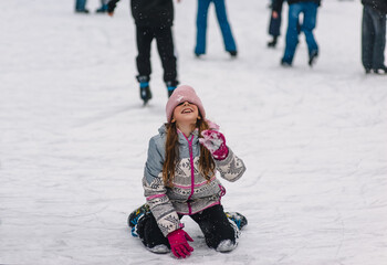 A beautiful happy teenage girl, child, lies in the snow on a snowy ice rink after slipping and falling outdoors in winter. Photography, portrait, sports, and leisure concept.