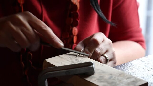 Close-up view of artisan hands filing a metal jewelry piece on a wooden bench pin. The craftsperson carefully shapes the object using a hand file in a bright workshop.

