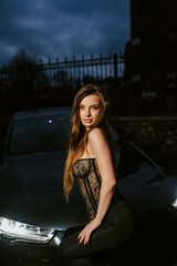 A photograph of a beautiful young sexy brunette woman, a fashionable model in lingerie, wearing a fur coat, standing at night in the dark with a luxurious expensive black car on a city street.