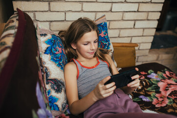 A beautiful girl, a teenage child, lies on a bed at home with a smartphone in her hands, watching a video, playing on a gadget.