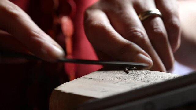 Close-up view of artisan hands filing a metal jewelry piece on a wooden bench pin. The craftsperson carefully shapes the object using a hand file in a bright workshop.

