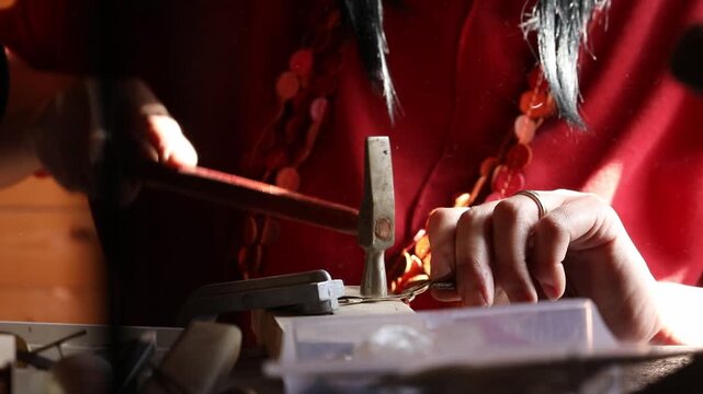 Close-up view of artisan hands filing a metal jewelry piece on a wooden bench pin. The craftsperson carefully shapes the object using a hand file in a bright workshop.

