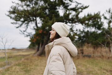 Fototapeta premium Outdoor scene with a person wearing a beige winter coat and knit beanie, standing in a park among trees and open field, calm weather and reflective mood.
