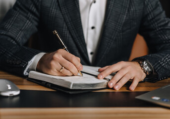 A close-up photo of an adult male businessman writing a business plan for a project in a notebook on a wooden desk in an office. Business concept.