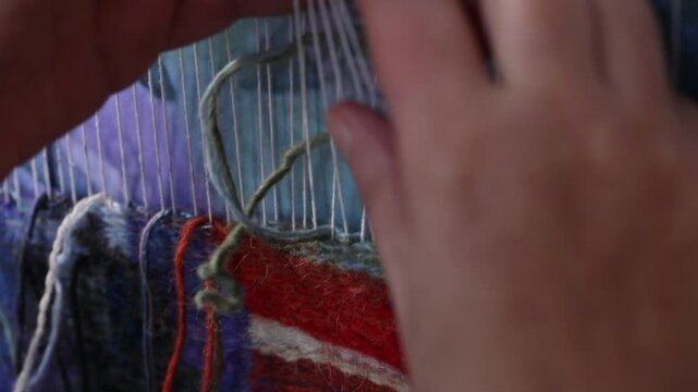 Close-up top view of artist hands weaving colorful tapestry on a loom. The artisan meticulously knots threads to create unique handmade textile wall art design.

